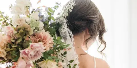 A bride from behind holding a large, romantic bouquet of pink and white flowers with greenery, wearing a floral hair accessory.