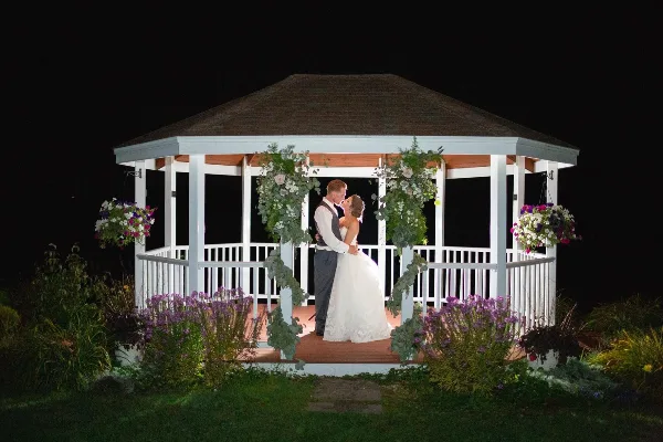 A bride and groom embracing in a white gazebo decorated with flowers at night.