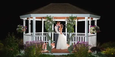 A bride and groom embracing in a white gazebo decorated with flowers at night.