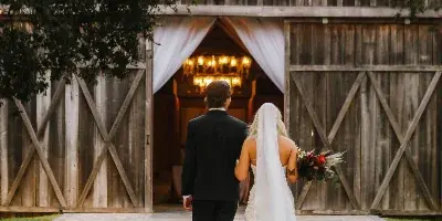 A bride and groom walking down a brick path toward a rustic wooden barn wedding venue decorated with string lights.