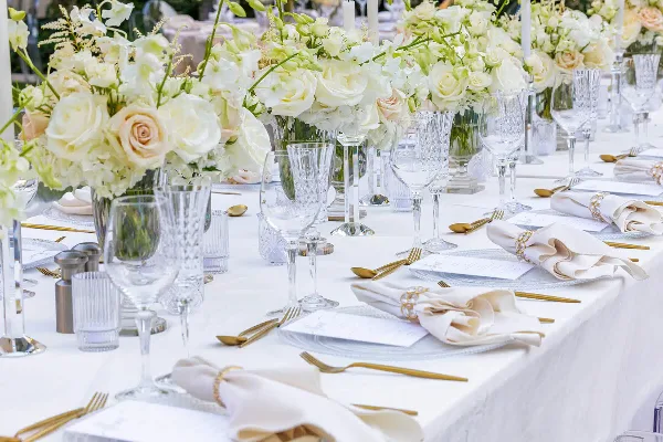 A professionally styled event table setting with white table linens, crystal glassware, gold flatware, and floral centerpieces.