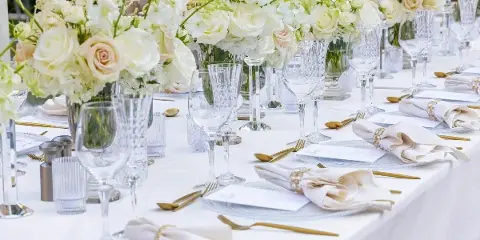 A professionally styled event table setting with white table linens, crystal glassware, gold flatware, and floral centerpieces.