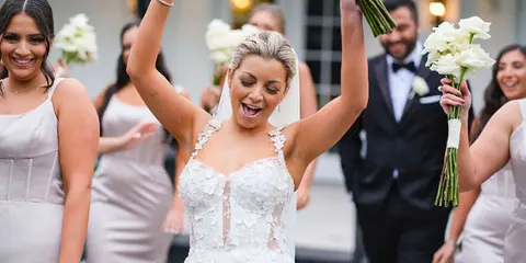 Bride cheers while holding her bouquet in the air as bridesmaids walk behind her