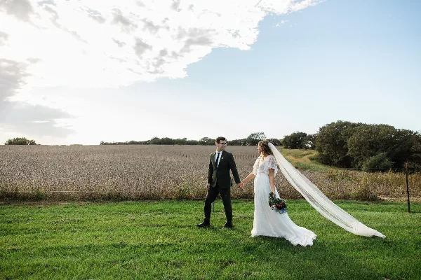 Bride and groom holding hands in a grassy field beside a crop field under a partly cloudy sky.