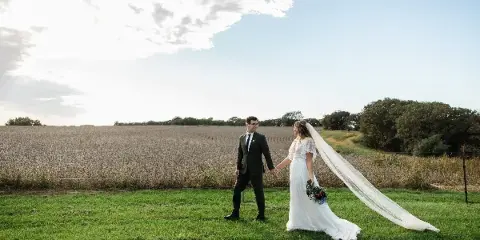 Bride and groom holding hands in a grassy field beside a crop field under a partly cloudy sky.