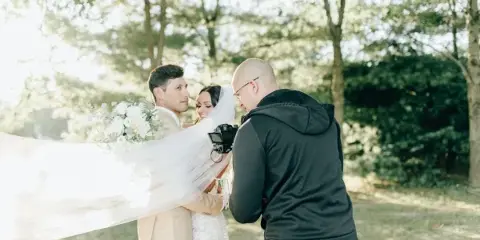 A videographer filming a bride and groom in an outdoor setting.