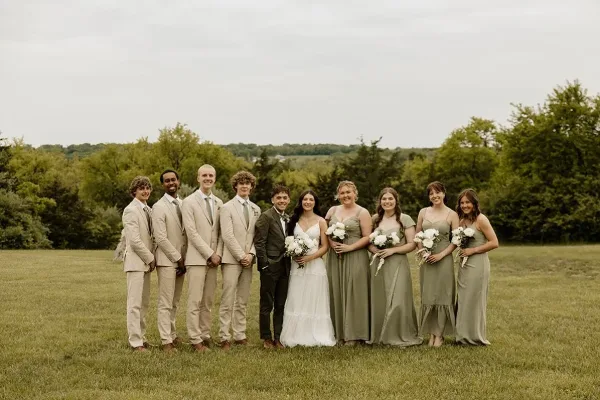 A wedding party consisting of a bride, groom, and attendants posing together in a grassy field with trees in the background.