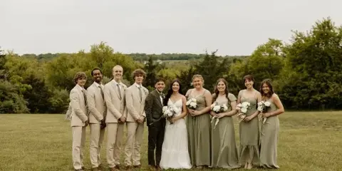 A wedding party consisting of a bride, groom, and attendants posing together in a grassy field with trees in the background.