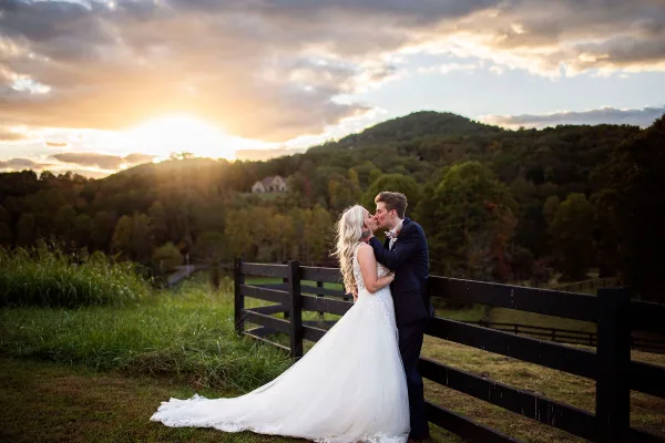 A bride and groom kissing in front of a wooden fence with a mountain landscape and sunset in the background.