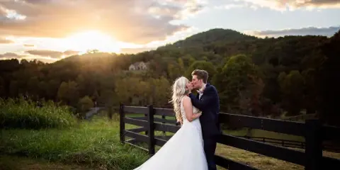 A bride and groom kissing in front of a wooden fence with a mountain landscape and sunset in the background.