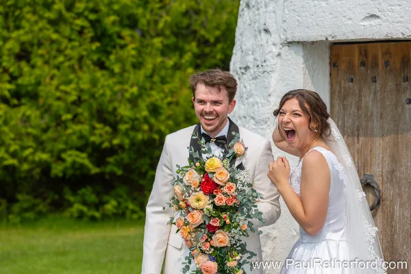 A bride and groom smiling and laughing outdoors, with the groom holding a large bouquet of flowers.
