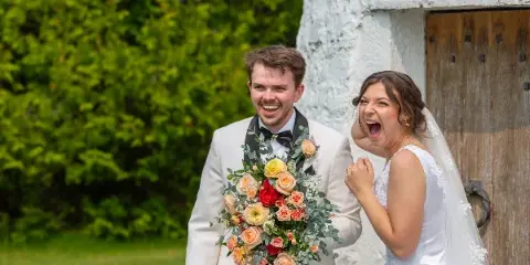 A bride and groom smiling and laughing outdoors, with the groom holding a large bouquet of flowers.