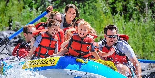 A group of smiling adults and children wearing life jackets ride a Timberline-branded raft on a river.