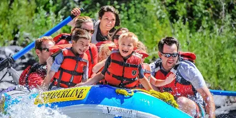 A group of smiling adults and children wearing life jackets ride a Timberline-branded raft on a river.