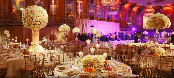 A large event venue set with round tables, white linens, chiavari chairs, floral centerpieces, and colored event lighting.