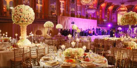 A large event venue set with round tables, white linens, chiavari chairs, floral centerpieces, and colored event lighting.