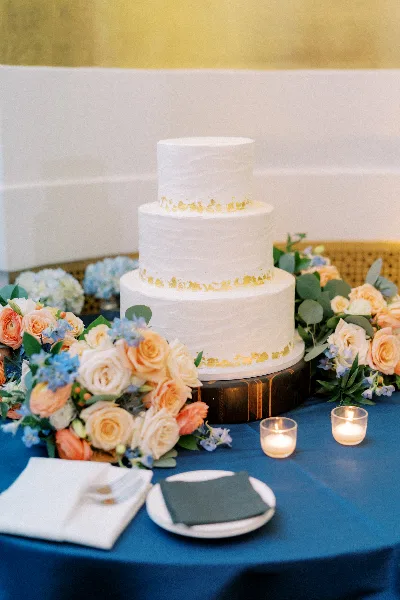 A three-tier white wedding cake with textured frosting and gold leaf accents, displayed on a wooden stand surrounded by floral arrangements and candles.