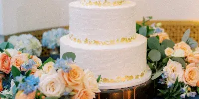 A three-tier white wedding cake with textured frosting and gold leaf accents, displayed on a wooden stand surrounded by floral arrangements and candles.