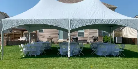 A white event tent set up in a residential backyard with round tables and chairs arranged underneath.