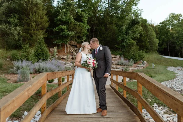 A bride and groom kissing on a wooden bridge surrounded by greenery and a stone wall.