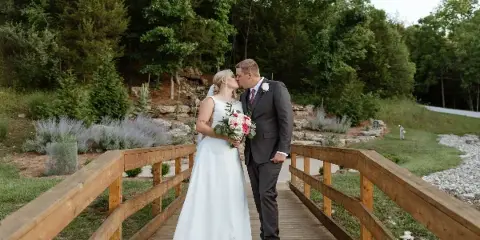 A bride and groom kissing on a wooden bridge surrounded by greenery and a stone wall.