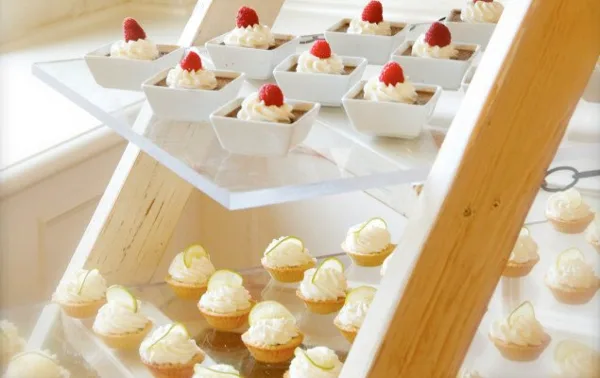 A tiered wooden display stand holding white square bowls of dessert topped with raspberries and small cupcakes with white frosting.
