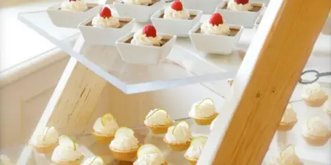 A tiered wooden display stand holding white square bowls of dessert topped with raspberries and small cupcakes with white frosting.