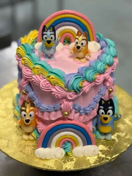 A custom two-tier cake decorated with colorful piped frosting, rainbow arches, and fondant figures of characters from the show Bluey.