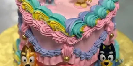 A custom two-tier cake decorated with colorful piped frosting, rainbow arches, and fondant figures of characters from the show Bluey.