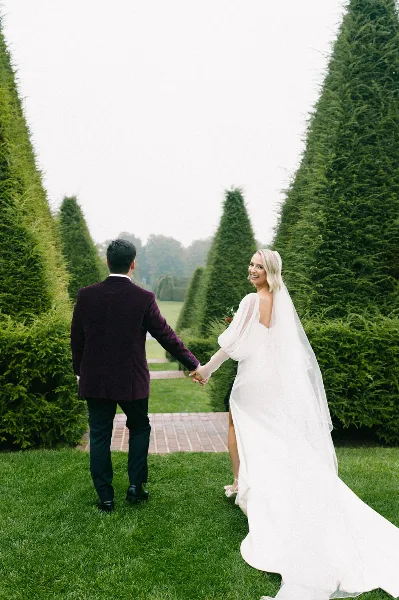 A bride and groom holding hands and walking through a manicured garden with tall conical trees.