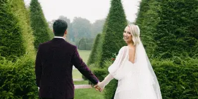 A bride and groom holding hands and walking through a manicured garden with tall conical trees.