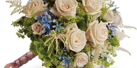 A round bouquet of cream roses and blue flowers with a pink ribbon tied to the stems.