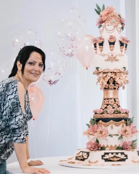 A woman posing next to a tall, ornate multi-tiered wedding cake decorated with pink flowers and gold accents.