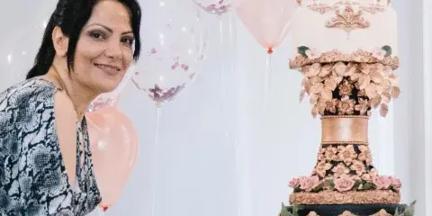 A woman posing next to a tall, ornate multi-tiered wedding cake decorated with pink flowers and gold accents.