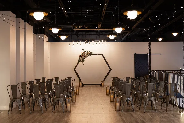 Indoor wedding ceremony setup with rows of metal chairs, aisle candles, hanging lights, and a floral-decorated hexagonal arch.