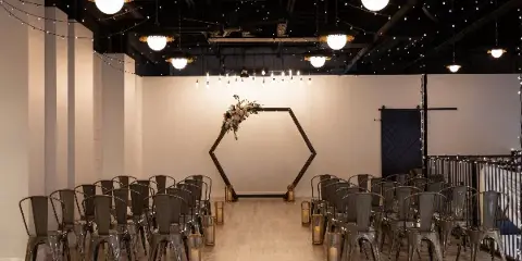 Indoor wedding ceremony setup with rows of metal chairs, aisle candles, hanging lights, and a floral-decorated hexagonal arch.