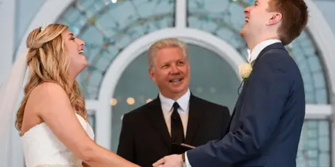 A bride and groom holding hands during a wedding ceremony with an officiant standing behind them.