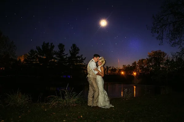 A bride and groom embracing at night by a pond with a full moon in the sky.