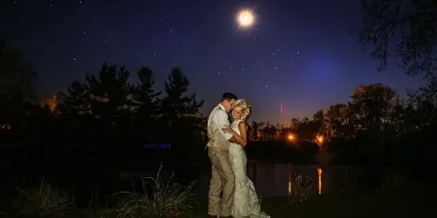 A bride and groom embracing at night by a pond with a full moon in the sky.