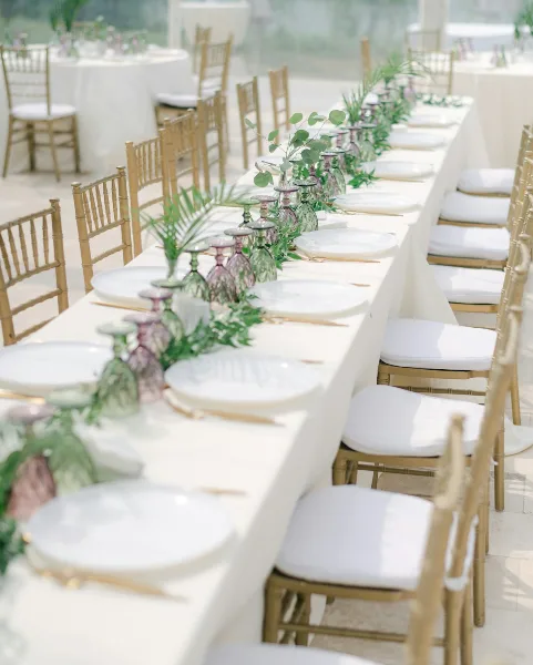 A long reception table set with white linens, gold Chiavari chairs, and a floral centerpiece.
