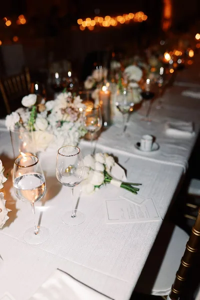 A close-up of an elegant wedding table setting featuring white floral arrangements, lit candles, and glassware on a white tablecloth.