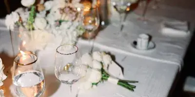 A close-up of an elegant wedding table setting featuring white floral arrangements, lit candles, and glassware on a white tablecloth.