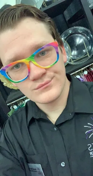A staff member wearing a black shirt and rainbow glasses stands in a showroom with shelves of silver chargers and hanging linens in the background.