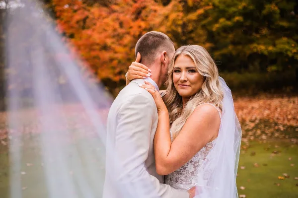 A bride and groom embracing outdoors with autumn foliage in the background.