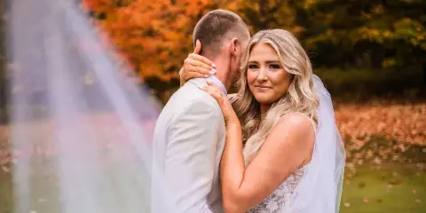 A bride and groom embracing outdoors with autumn foliage in the background.