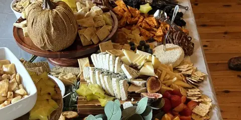 A large, elaborately arranged catering board featuring various cheeses, crackers, dried fruits, and a decorative pumpkin centerpiece.