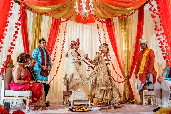 A bride and groom performing a wedding ritual in front of a decorated red and gold mandap with floral garlands.