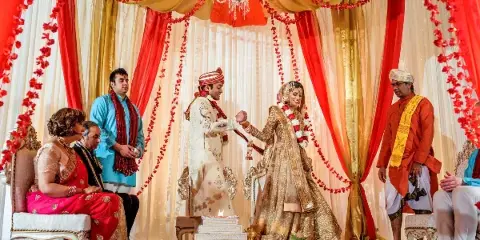 A bride and groom performing a wedding ritual in front of a decorated red and gold mandap with floral garlands.