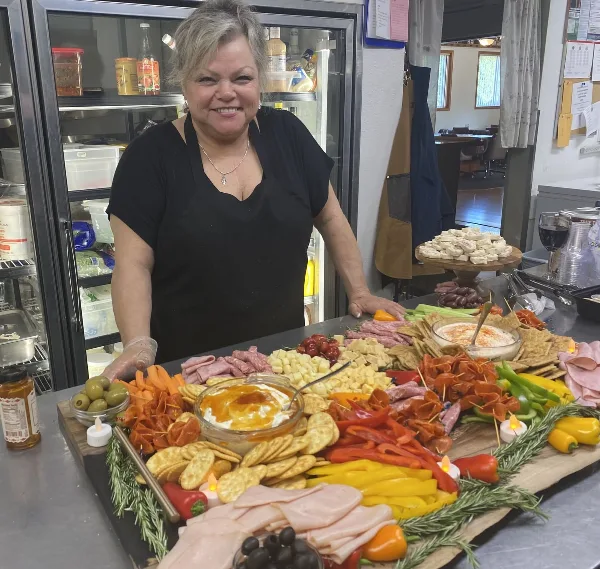 A smiling woman in a black apron stands behind a large, colorful catering spread featuring meats, cheeses, vegetables, and dips on a wooden board.