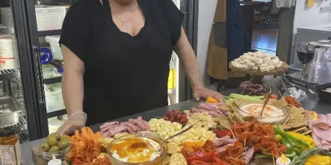 A smiling woman in a black apron stands behind a large, colorful catering spread featuring meats, cheeses, vegetables, and dips on a wooden board.